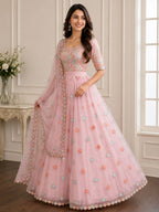 A woman models a light pink traditional Indian anarkali gown with a heavily embroidered peach and blue bodice, a matching pink waistband, and a full tulle skirt decorated with embroidered floral motifs and a sheer dupatta.