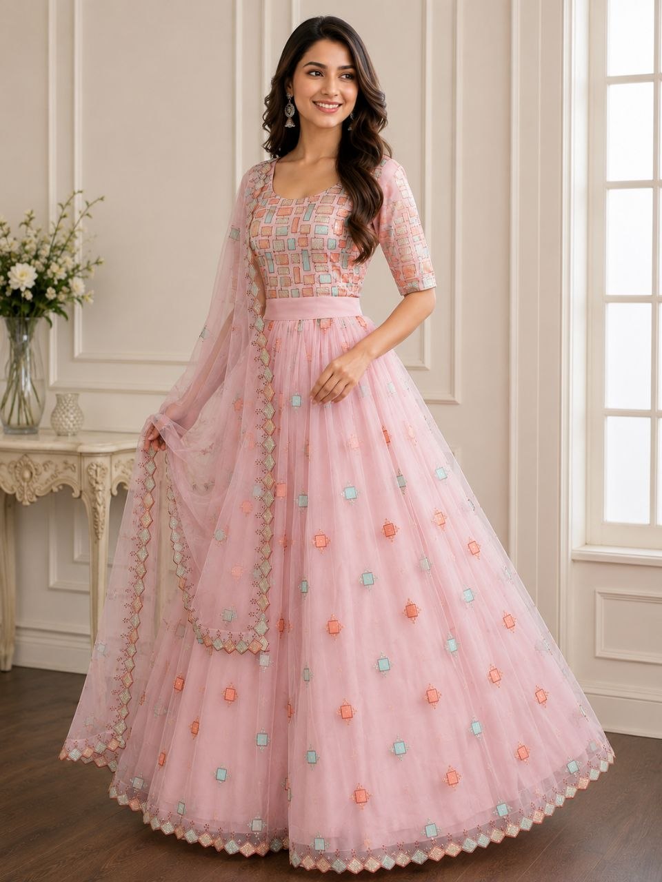 A woman models a light pink traditional Indian anarkali gown with a heavily embroidered peach and blue bodice, a matching pink waistband, and a full tulle skirt decorated with embroidered floral motifs and a sheer dupatta.