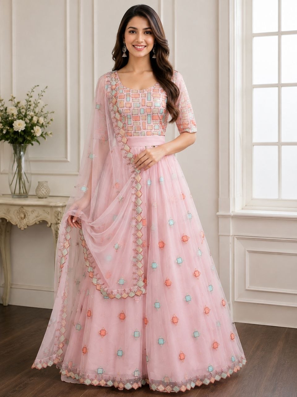 A woman models a light pink traditional Indian anarkali gown with a matching dupatta in a bright, elegant room. The dress features a heavily embroidered geometric patterned bodice, short sleeves, and a flared skirt decorated with small floral motifs in pastel orange, green, and blue.