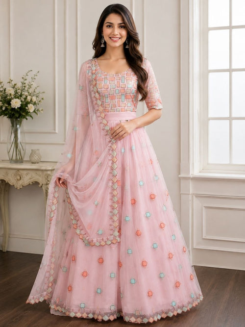 A woman models a light pink traditional Indian anarkali gown with a matching dupatta in a bright, elegant room. The dress features a heavily embroidered geometric patterned bodice, short sleeves, and a flared skirt decorated with small floral motifs in pastel orange, green, and blue.